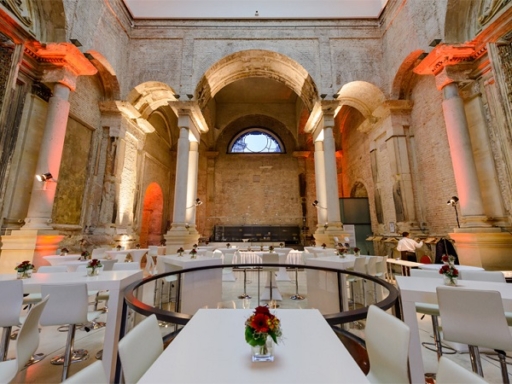 Im Lipsiusbau der Technischen Universität Dresden ist ein Bankett vorbereitet. Weiße Tische und weiße Stühle mit Blumen darauf stehen bereit. Ein Mann richtet mehrere Lampen ein.