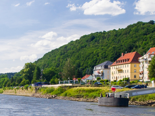 A steamer sails upstream on the Elbe River in Saxon Switzerland. The ship is about to reach the pier in Bad Schandau. The Elbe bridges in Bad Schandau and the Lilienstein mountain can be seen in the background. It's sunny with a few clouds.