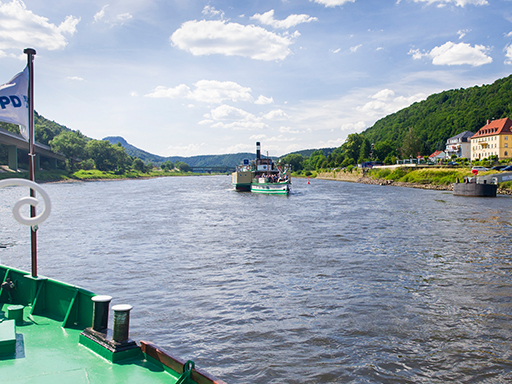 Ein Dampfer fährt auf der Elbe in der Sächsischen Schweiz stromauf. Gleich erreicht das Schiff die Anlegestelle in Bad Schandau. Im Hintergrund sind die Elbbrücken in Bad Schandau und der Lilienstein zu sehen. Es ist sonnig mit ein paar Wölkchen. Im Vordergrund ist der Bug eines weiteren Dampfers zu sehen, von dem aus das Foto geschossen wurde.