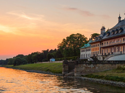 Evening castle tour A steamer sails upstream past Pillnitz Castle. Many people sit on the upper deck. It's early evening, and the setting sun bathes the scenery in an orange glow.