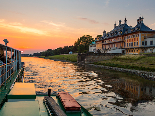 Ein Dampfer fährt stromauf an Schloss Pillnitz vorbei. Auf dem Oberdeck sitzen viele Menschen. Es ist früher Abend, die untergehende Sonne taucht die Szenerie in orangefarbenes Licht.