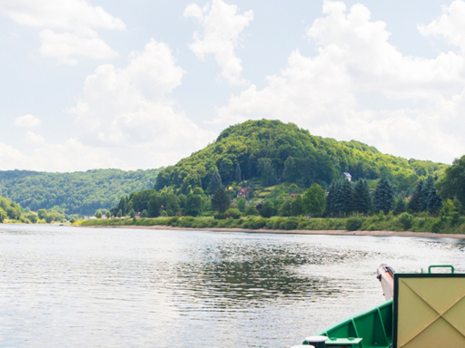 Ein Dampfer fährt elbaufwärts durch die Sächsische Schweiz. Die bewaldeten Elbufer sind zu sehen, im Vordergrund die goldene Schiffsglocke.