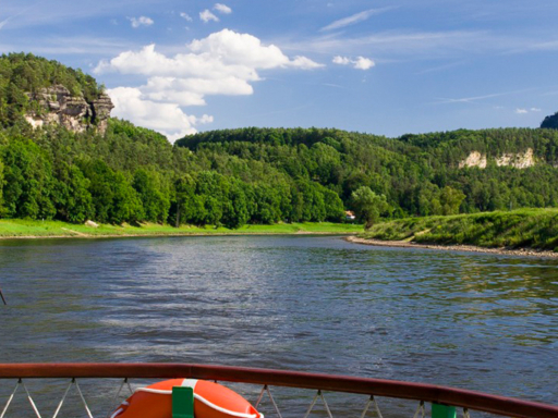 View from the stern of a steamer sailing downstream in Saxon Switzerland. Rocky cliffs and Königstein Fortress can be seen in the background. It's summer, and the trees and meadows are green.