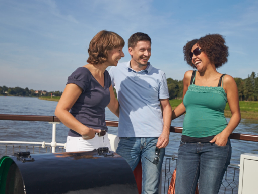 Auf einem Schiff stehen zwei Frauen und ein Mann an der Reling. Sie lachen, unterhalten sich und genießen die Fahrt bei Sonnenschein und wolkenlosem Himmel.