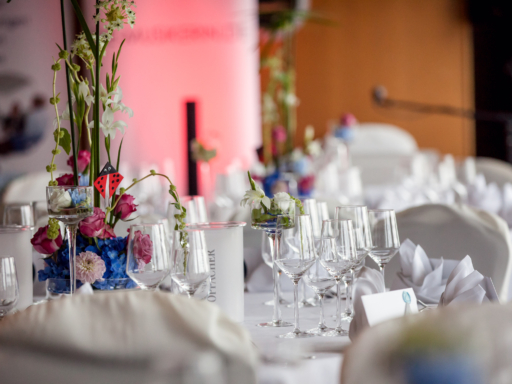 Auf einem Tisch stehen Gläser bereit, Servietten sind aufgestellt, ein Blumengesteck mit rosafarbenen Rosen schmückt den Tisch.