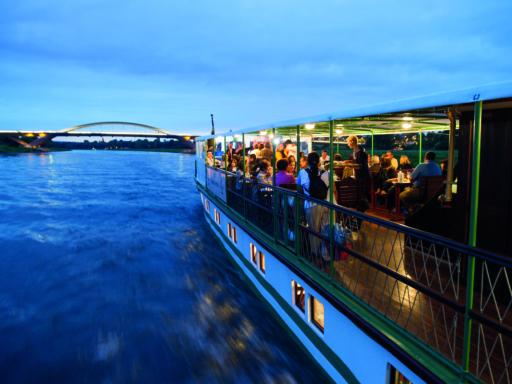 Fahrgäste sitzen abends an den Tischen auf einem Dampfer, der auf der Elbe stromauf gerade die Dresdner Waldschlößchenbrücke passiert hat.