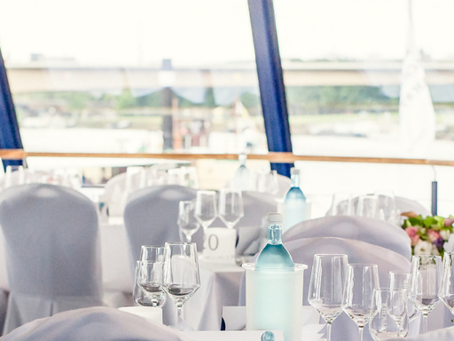 On the upper deck of a saloon ship, tables and chairs are set for a festive event. White covers are placed over the chairs, and the tables are set. Flowers are placed on the tables.