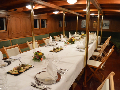 In the lower deck saloon of a steamer, a long table is set. Plates, glasses, and cutlery are prepared, and flowers and appetizers are on the table.