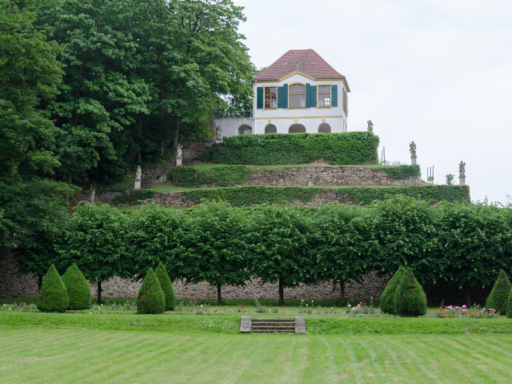 Die Heinrichsburg in Seußlitz. Das zweistöckige barocke Gartenhaus steht auf einer Anhöhe am südlichen Ende des Schlossparks Seußlitz. Im Vordergrund sind spitz beschnittene Büsche zu sehen.