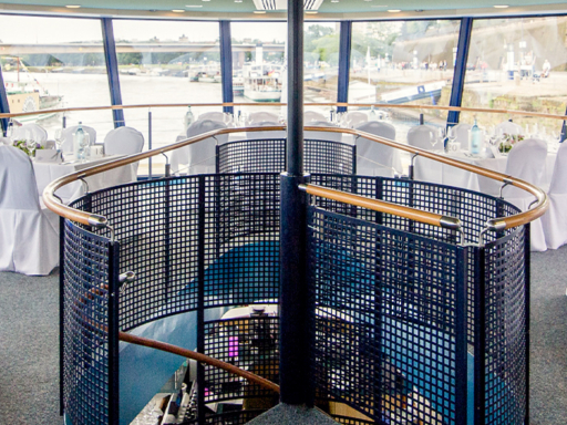 On the upper deck of a saloon ship, tables and chairs are set for a festive event. White covers are placed over the chairs, and the tables are set. Flowers are placed on the tables.