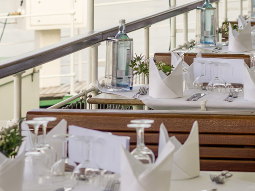Tables are set on the upper deck of a steamer. Glasses are on the tables, cutlery is ready, and water bottles are in coolers.