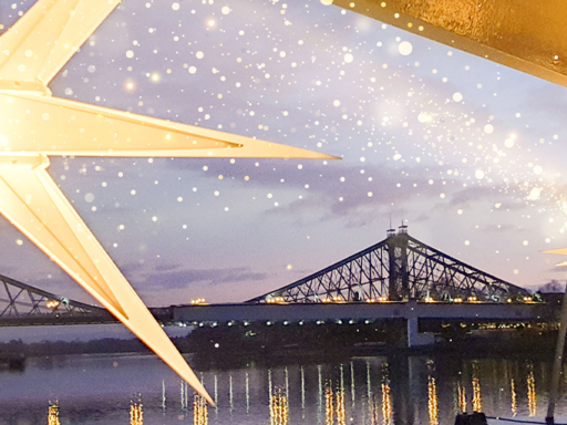 Zwei weiße Herrnhuter Adventssterne hängen auf einem Schiff, das abends auf das Blaue Wunder zufährt.