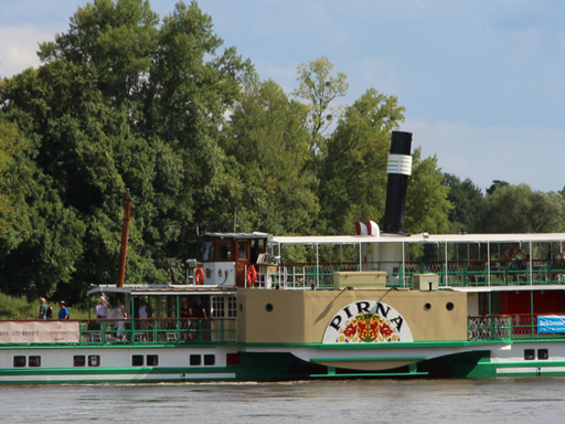 Header_0002_PD Pirna The steamer Pirna sails into town on a sunny summer day. The ship's chimney is up, the sky is blue, and the trees and meadows are green.