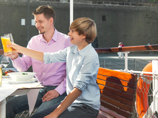 Eine Familie mit zwei Kinder sitzt bei schönem Wetter an Deck eines Schiffes und stößt mit Softdrinks an. Auf einem Tisch stehen Teller mit Essen darauf.