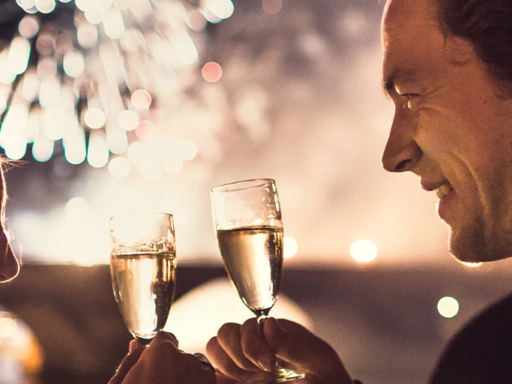 Ein Pärchen stößt mit Sekt an, im Hintergrund ist es dunkel, nur Feuerwerk ist zu erkennen.