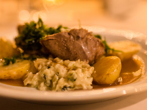 Auf einem eingedeckten Tisch stehen im Hintergrund mehrere Weingläser, eins davon ist mit Rotwein gefüllt. Auf einem Teller im Vordergrund liegt ein Stück Gänsebraten mit Soße, Gemüse und Kartoffeln.
