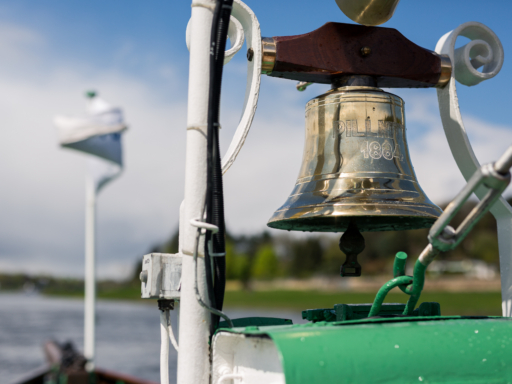 Auf einem Schiff hängt die goldene Schiffsglocke. Im Hintergrund sind verschwommen der Fluss und grüne Bäume zu sehen.