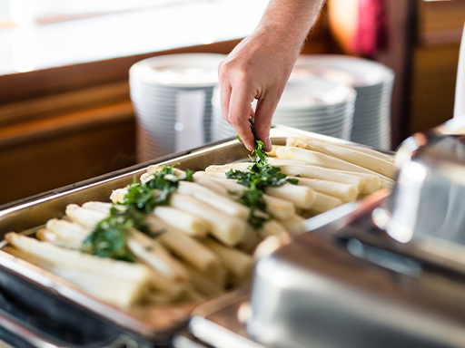 Speisen In einem Warmhalte-Behälter liegt Spargel. Ein Mitarbeiter bestreut ihn mit Petersilie.