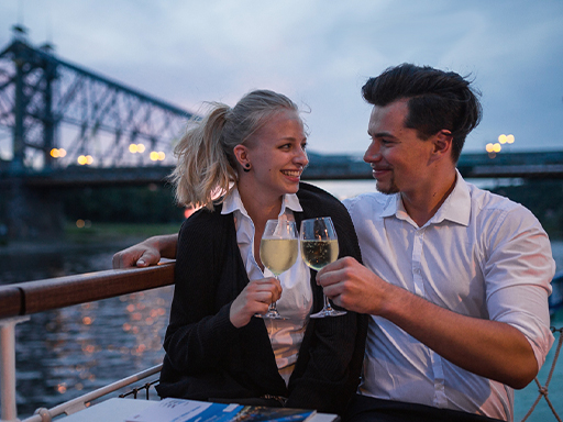 Ein Pärchen stößt auf einem Schiff mit Wein an. Das Schiff hat gerade das Blaue Wunder erreicht, es ist Abend.