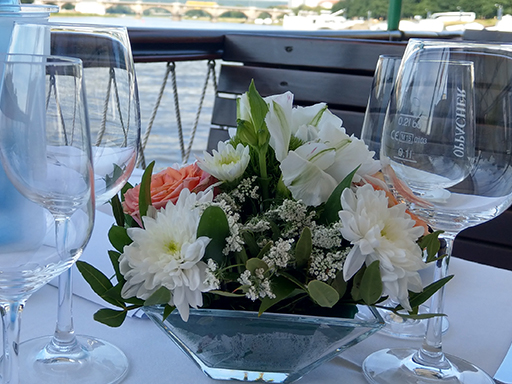Auf einem Tisch auf dem Oberdeck eines Dampfers stehen Gläser und ein Blumengesteck mit weißen und rosafarbenen Blüten.