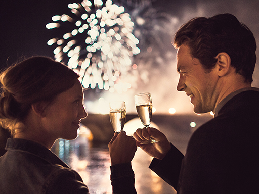 Ein Pärchen stößt mit Sekt an. Dahinter ist verschwommen eine Brücke zu sehen, am Himmel Feuerwerk.
