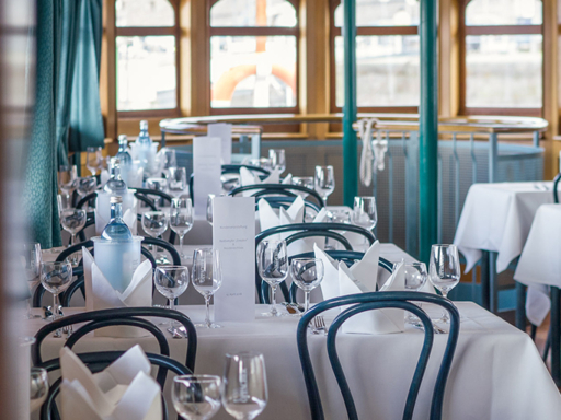 In the deck saloon of a steamer, all tables are set with white tablecloths, plates, glasses, cutlery and napkins.