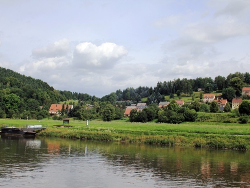 Die Anlegestelle an der Elbwiese in Prossen. Im Hintergrund stehen die Häuser des Dorfs, die Bäume sind grün, am blauen Himmel sind ein paar weiße Wolken zu sehen.