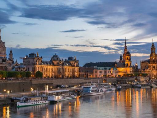 Die Silhouette der Dresdner Altstadt in der Abenddämmerung mit Blick auf das Terrassenufer, wo mehrere Dampfer und Salonschiffe anliegen. Die Lichter der Altstadt spiegeln sich im Wasser der Elbe.