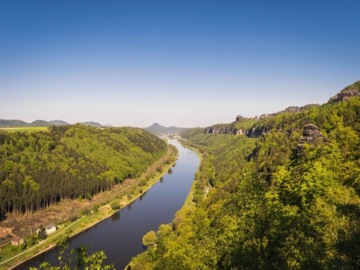 Blick auf die Elbe stromab in Richtung Lilienstein. Die Elbhänge sind grün, der Himmel blau, es sind keine Wolken zu sehen. Links verläuft die Bahnstrecke nach Dresden, aber es fährt gerade kein Zug.