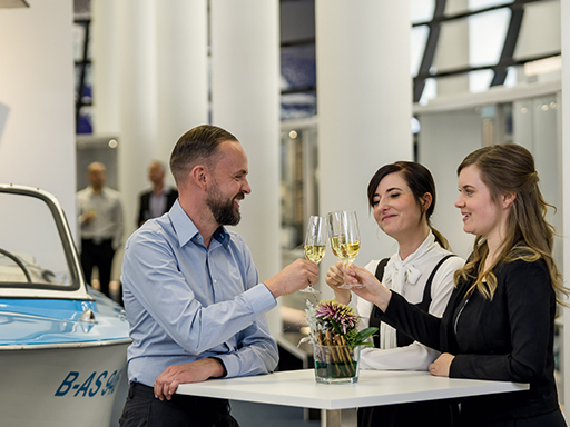 Zwei Frauen und ein Mann stehen an einem Stehtisch und stoßen mit Sektgläsern an.