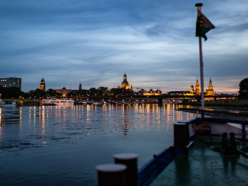 Ein Dampfer fährt stromab und hat gerade die Albertbrücke passiert. Gleich erreicht er das beleuchtete Stadtzentrum. Es ist Abend, im Hintergrund funkeln die Lichter der Stadt.