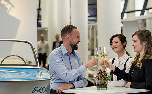 An einem Stehtisch stoßen zwei Frauen und ein Mann mit Weingläsern an.