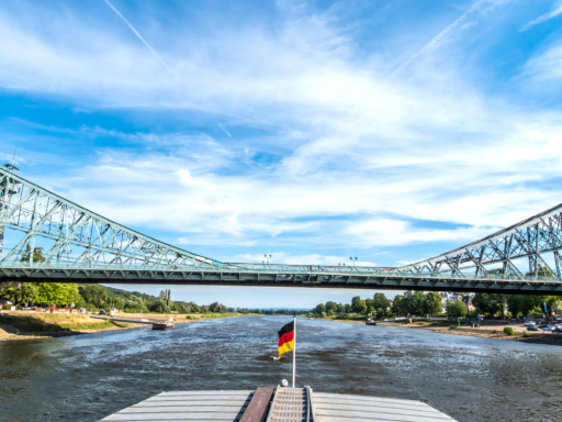 Ein Dampfer fährt auf der Elbe stromab. Er hat gerade das Blaue Wunder passiert, zeigt der Blick zum Heck des Schiffs. Es ist sonnig, aber windig.