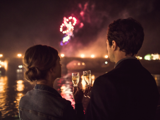 Ein Pärchen stößt mit Sekt an. Dahinter ist verschwommen eine Brücke zu sehen, am Himmel Feuerwerk.