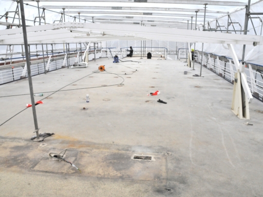 2024-02-27-Umbau Cosel aktueller Stand (3) The sun deck of a motor ship. It's empty; at the very back, a man is kneeling and working. The ship is covered in scaffolding, tarpaulins making the structure look like a closed garage.