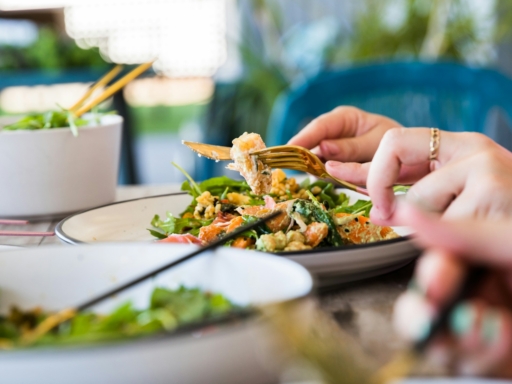 Auf einem Tisch stehen mehrere Teller mit Salaten. Eine Person isst von einem der Teller, die Hände, Messer und Gabel sind zu sehen.