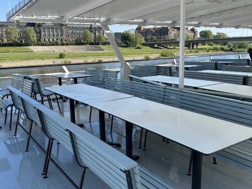 image00013 Mintfarbene Bänke und weiße Tische stehen auf dem Sonnendeck eines Motorschiffs. Das Schiff liegt am Terrassenufer, von oben ist die Elbe zu sehen.