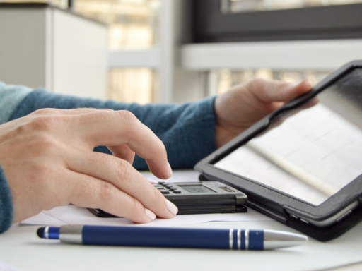 Buchhaltung Auf einem Tisch liegt ein Taschenrechner, davor ein blauer Kuli. Eine Person liest Zahlen von einem Bildschirm ab und tippt sie in den Taschenrechner.