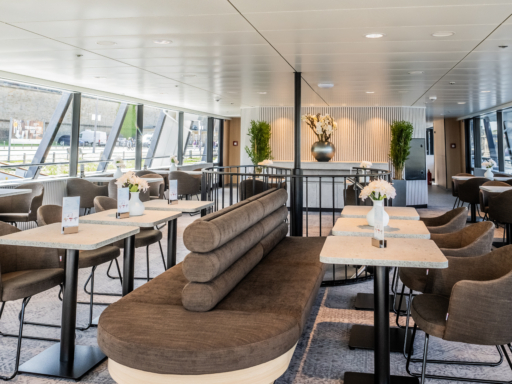 Blick von vorn in den Fine Dining Club im Oberdeck eines Salonschiffs. Braune Sitzmöbel und helle Tische bestimmen das Bild, im Hintergrund ein Tresen mit weißen Blumen.