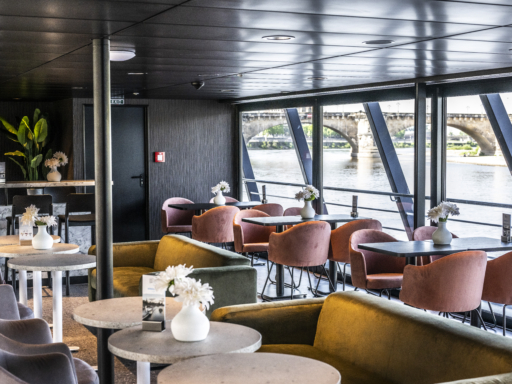 Blick durch das Lounge Café eines Salonschiffs auf die Elbe. Im Vordergrund sind gemütliche Sofas und Sitze im Café zu sehen, dunkle, warme Farben bestimmen das Ambiente. Auf den Tischen stehen weiße Blumen. Im Hintergrund die Elbe mit der Augustusbrücke.