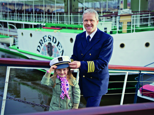 The captain of the steamer Dresden, Andreas Weber, stands on the bridge in uniform. He places his captain's hat on the head of a little girl, about six years old. Both smile at the camera.
