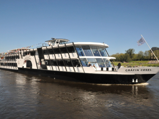 Das Salonschiff Gräfin Cosel legt geschmückt mit bunten Wimpeln vom Terrassenufer ab. Auf der Wiese am Königsufer stehen viele Leute.