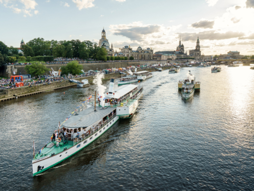 Sechs Dampfer fahren mit bunten Wimpeln geschmückt am Terrassenufer ab. Bei den vorderen drei Dampfern steigt Dampf aus der Pfeife auf. Es sind große Menschenmengen am Terrassenufer und auf der Brühlschen Terrasse unterwegs. Das Wetter ist sonnig mit einzelnen Wolken am Himmel.