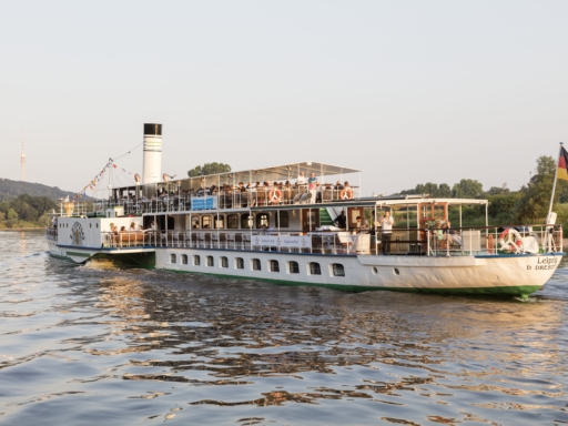 Der Dampfer Leipzig fährt an einem sonnigen Tag die Elbe Richtung Blasewitz entlang. In der Ferne erkennt man den Fernsehturm. Das Schiff ist voller Menschen. Am Heck weht eine Deutschlandfahne.