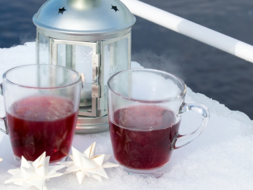 Zwei Teegläser mit rotem Glühwein gefüllt stehen auf einem schneebedeckten Tisch auf dem Sonnendeck eines Schiffes, umgeben werden diese von drei gebastelten Sternen, im Hintergrund steht eine kleine Laterne aus Metall
