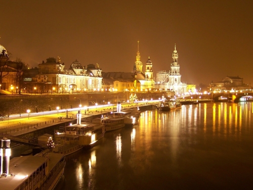 Three ships lie on the illuminated terraced bank in front of the city's silhouette on a snowy winter evening.
