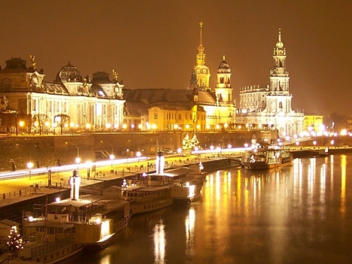Drei Schiffe liegen an einem Winterabend mit Schnee am beleuchteten Terrassenufer vor der Silhouette der Stadt.